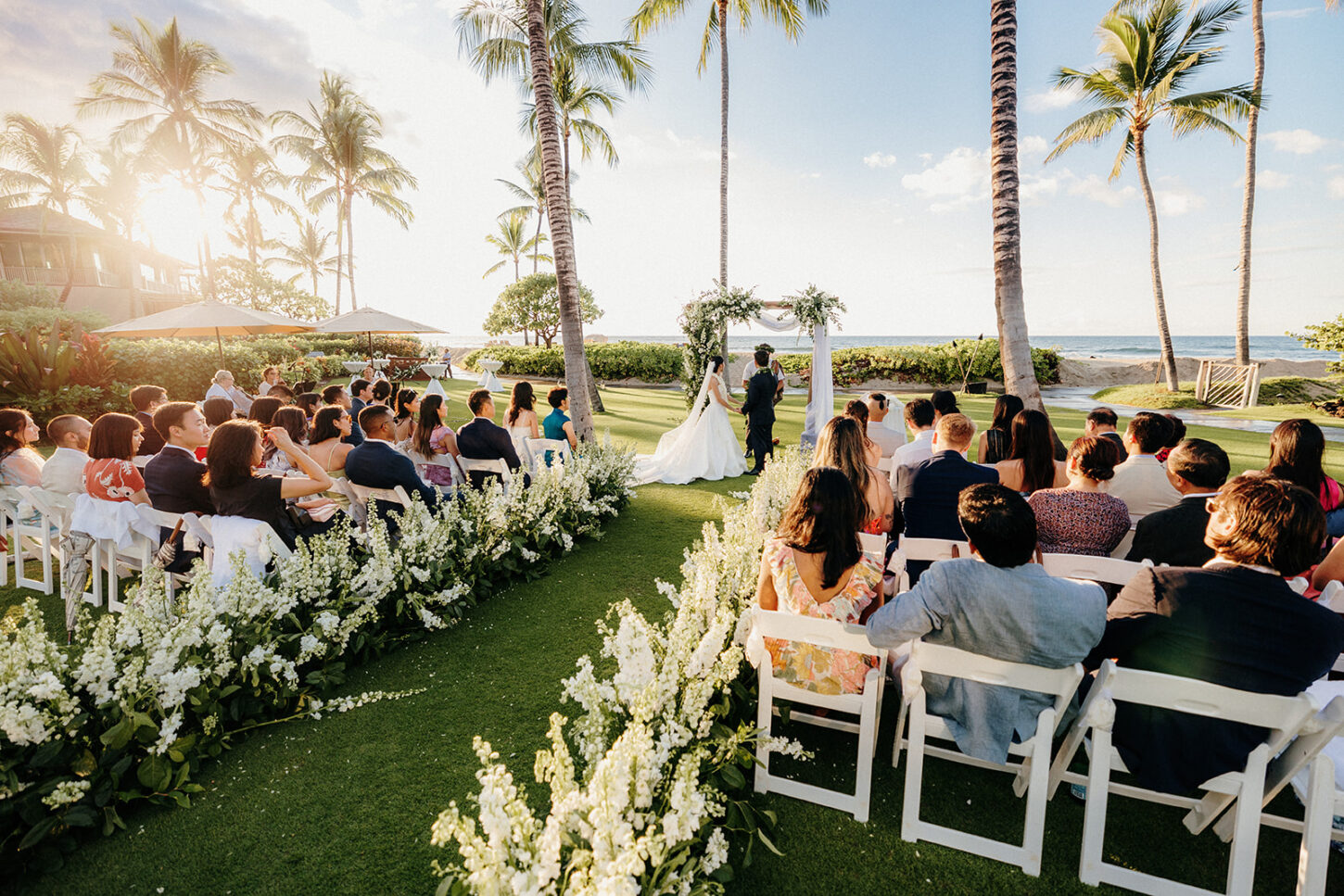 A bride and groom exchanging vows under a floral arch, with lush greenery in the background and the ocean in the distance.